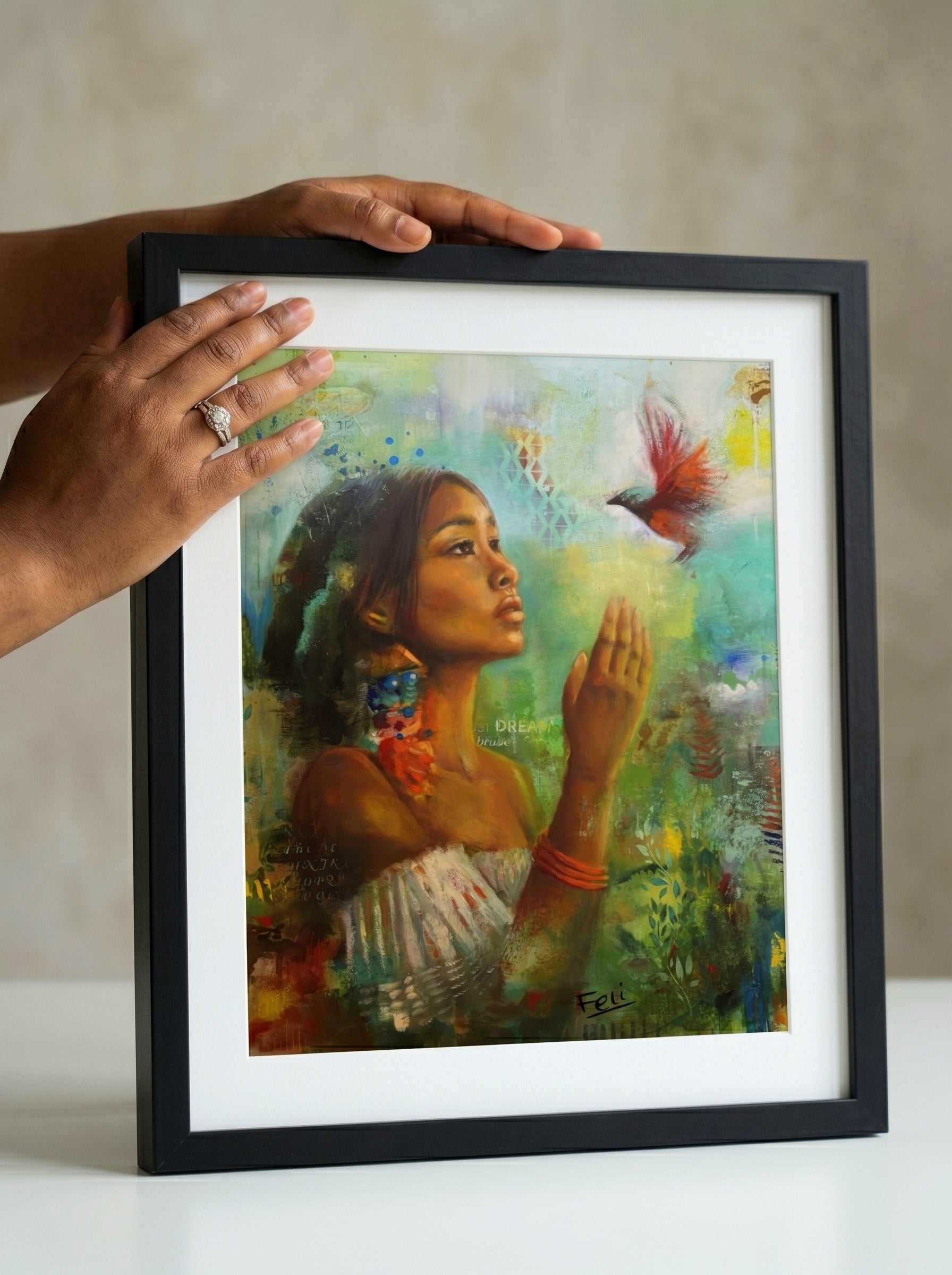 A framed art print entitled if i had wings by feli the artist featuring a colorful painting of a indian mixed black woman reaching for a red bird, held by two hands in a black picture frame against a neutral beige background.
