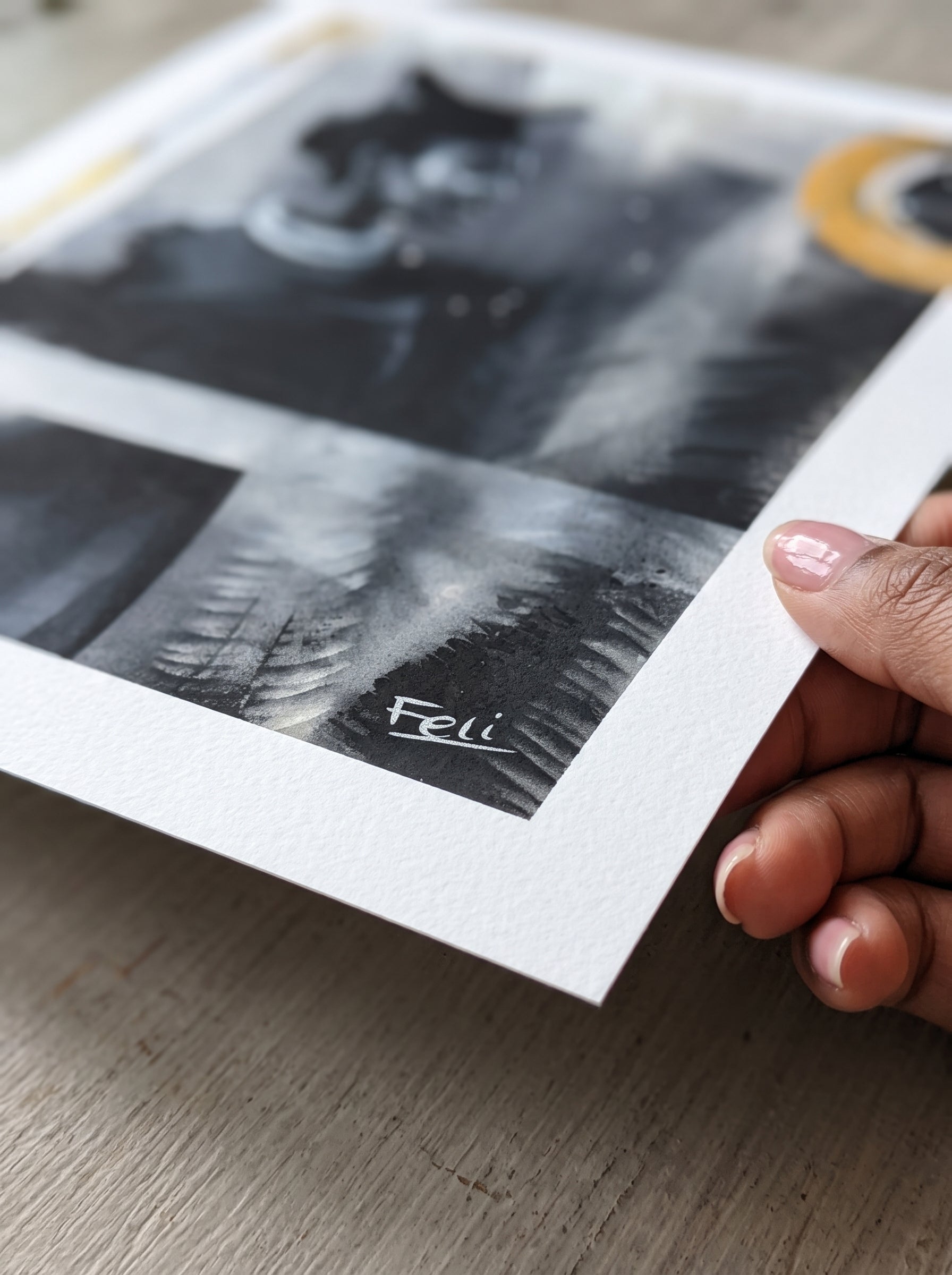 A close-up of a hand holding a printed art print over a light wood surface. The print features black-and-white abstract realism of a blurred out girl  in grey scale with yellow gold accents around and fossil impressions and this is by feli the artist entitled my footprints.

