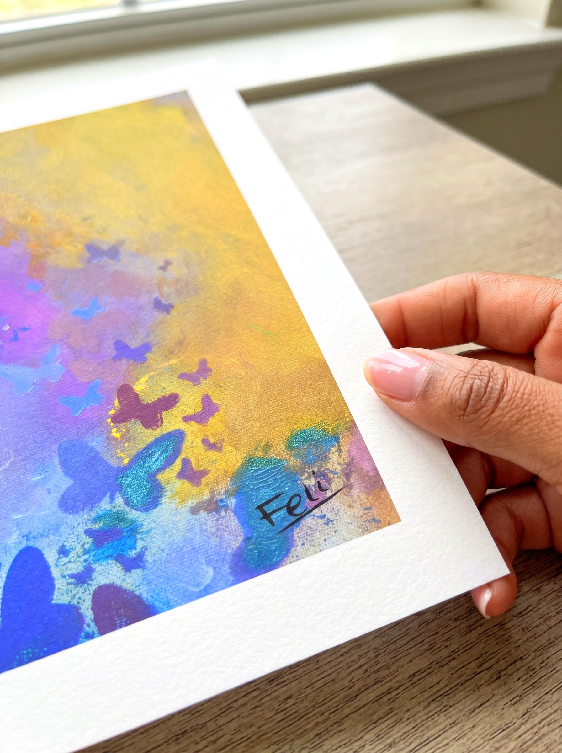 A hand holding the edge of a textured art print with a colorful abstract painting on it, featuring yellow, blue, purple, butterflies and teal tones which is entitled letting go by feli the artist.