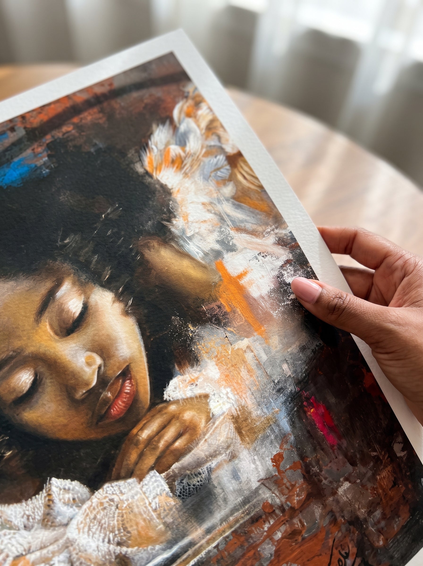 A hand holds a printed artwork entitled reflections by feli the artist showing a painted portrait of a black african girl woman angel with closed eyes, rendered in dark brown, black, white, and orange tones. The print is photographed over a softly blurred indoor background.
