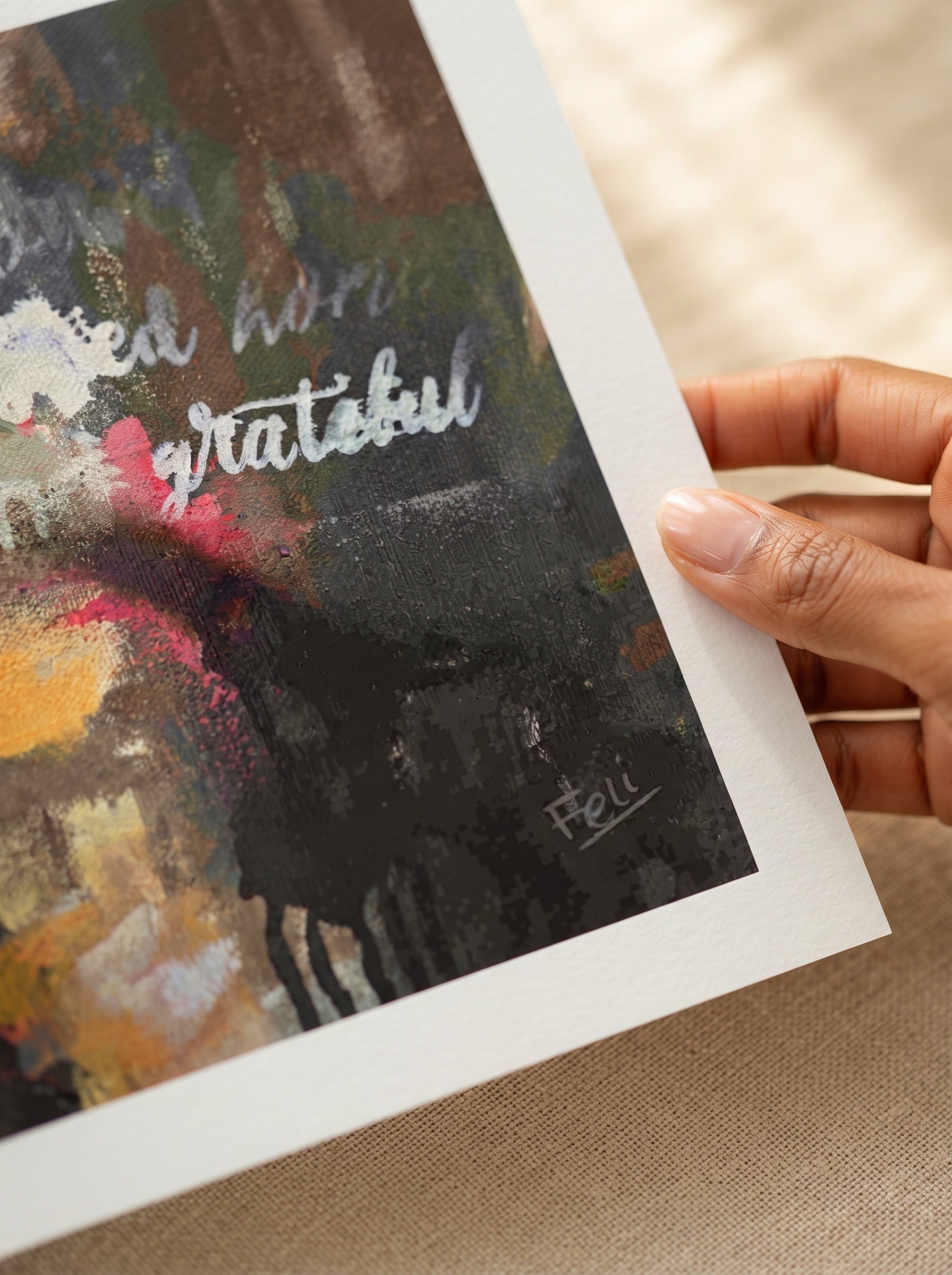 A close-up of a hand holding the edge of an art print titled grateful and blessed by feli the artist featuring an abstract painting with the words 'grateful' in white script over a dark, textured background.