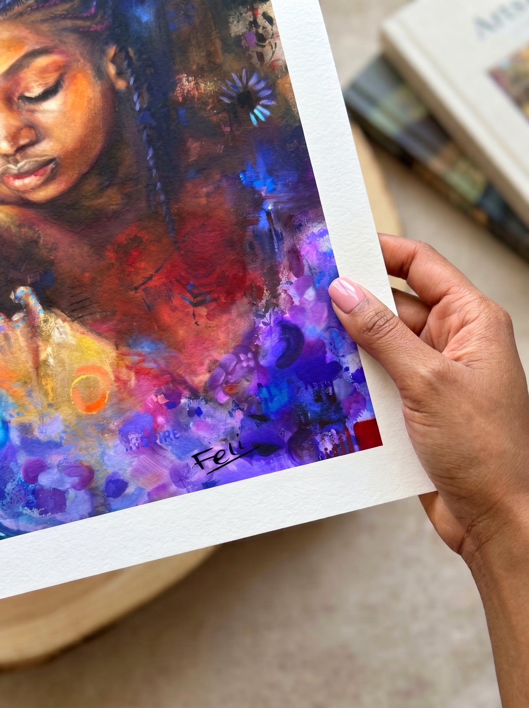 A hand holding a printed art piece entitled as she blooms by feli the artist featuring an abstract realism art print with rich purple, blue, red, and orange tones, set against a softly blurred indoor background.