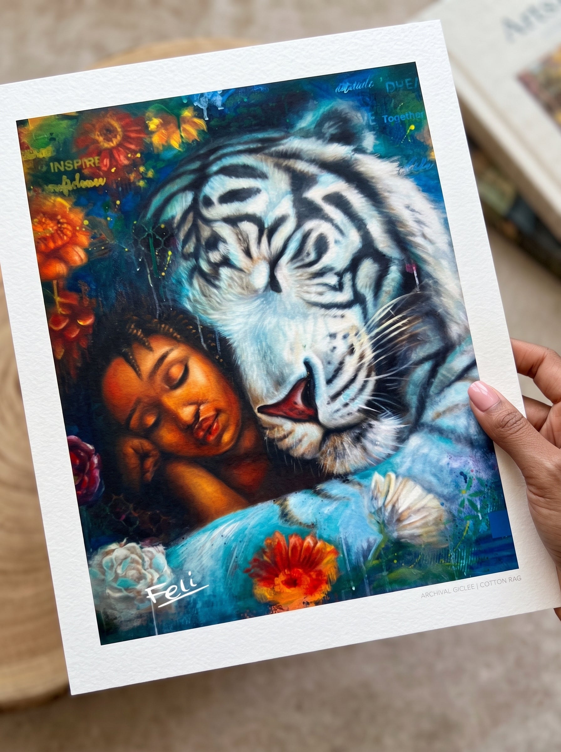 A hand holds a matted art print entitled wild heart gentle soul by feli the artist featuring a colorful illustration of a white tiger and a sleeping woman surrounded by flowers.
