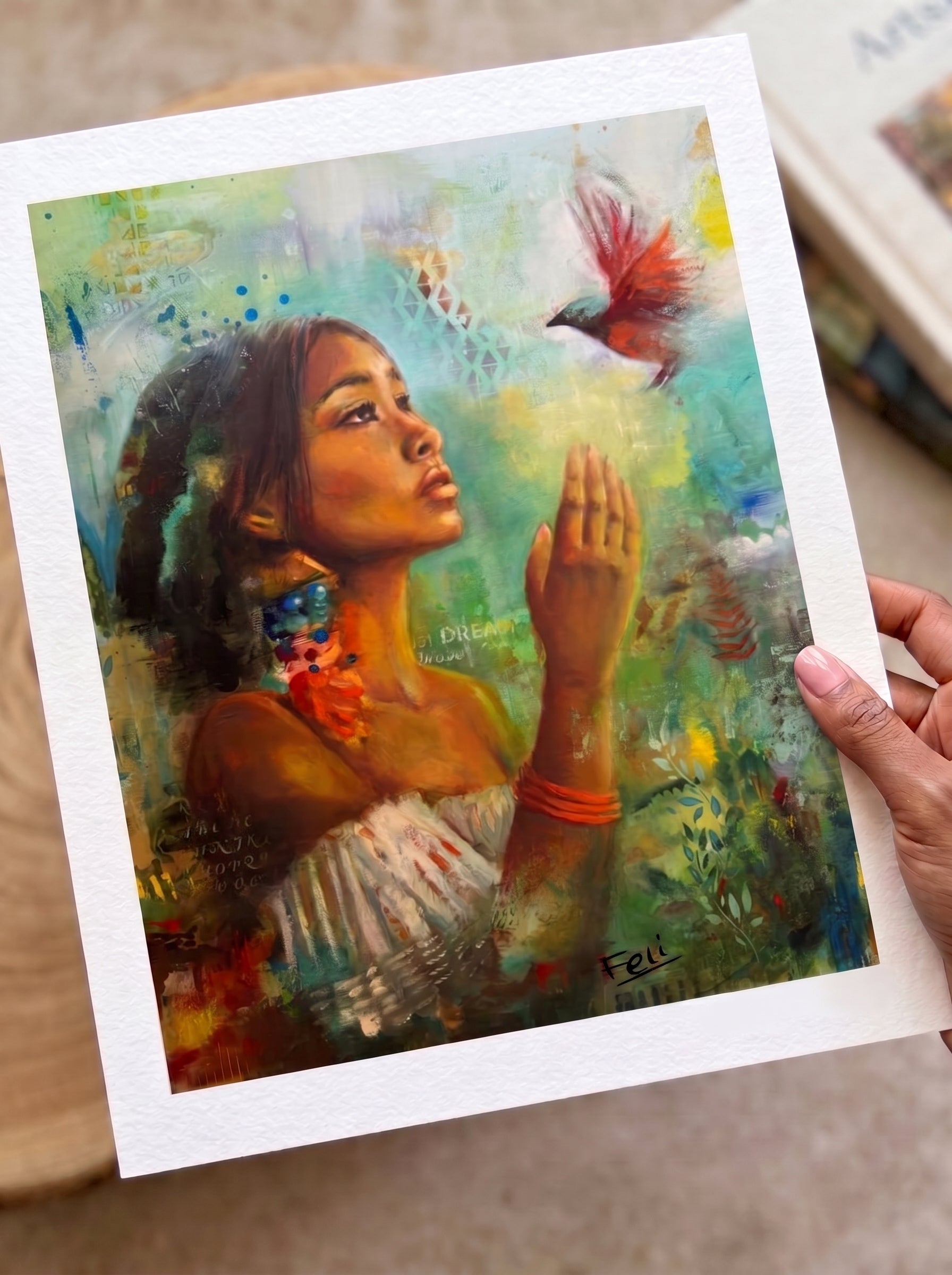 a hand holding the edge of An art print entiled if i had wings by feli the artist featuring a green floral colorful portrait of a indian black mixed woman reaching for a hummingbird, styled flat-lay on a white surface with scattered white and gold flowers.