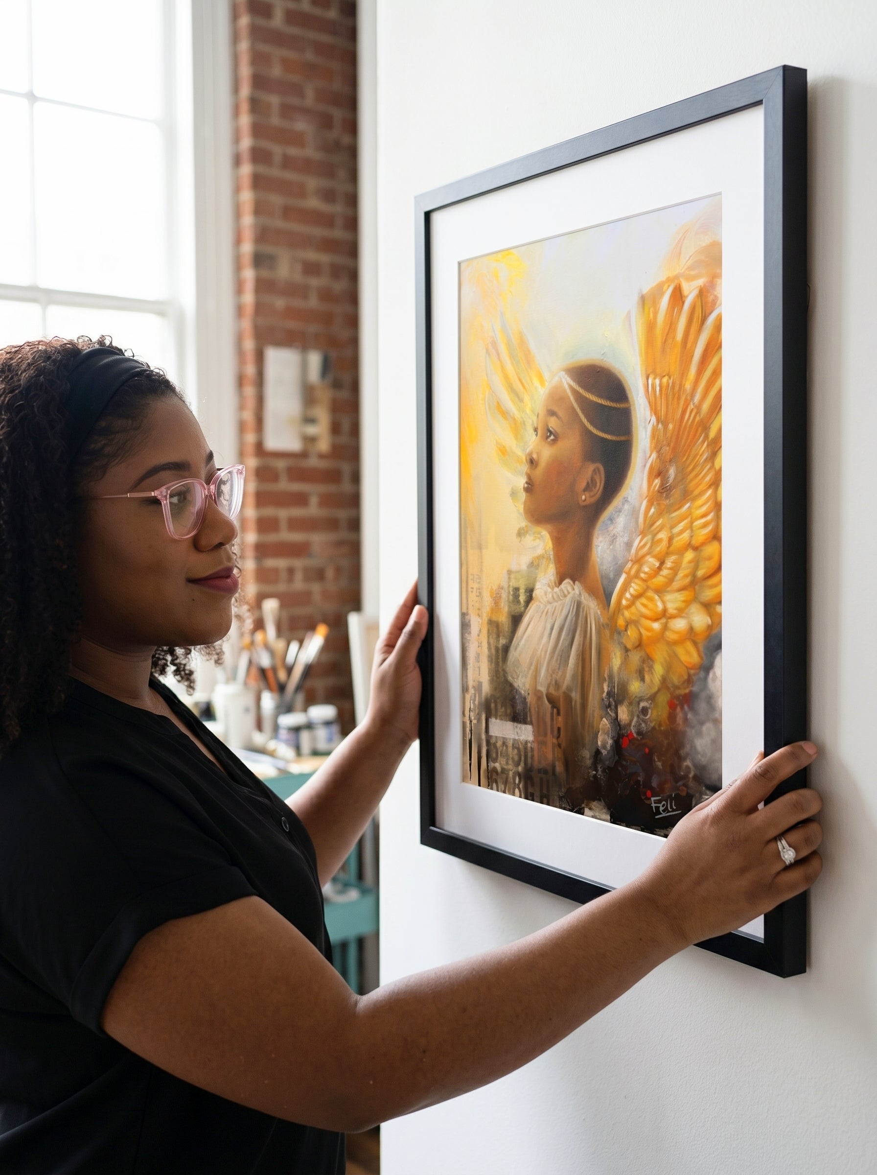 A woman in a black shirt and glasses hangs a large framed art print  featuring a black angel with gold accents in a black picture frame hang on a white wall in a bright room with a brick column.
