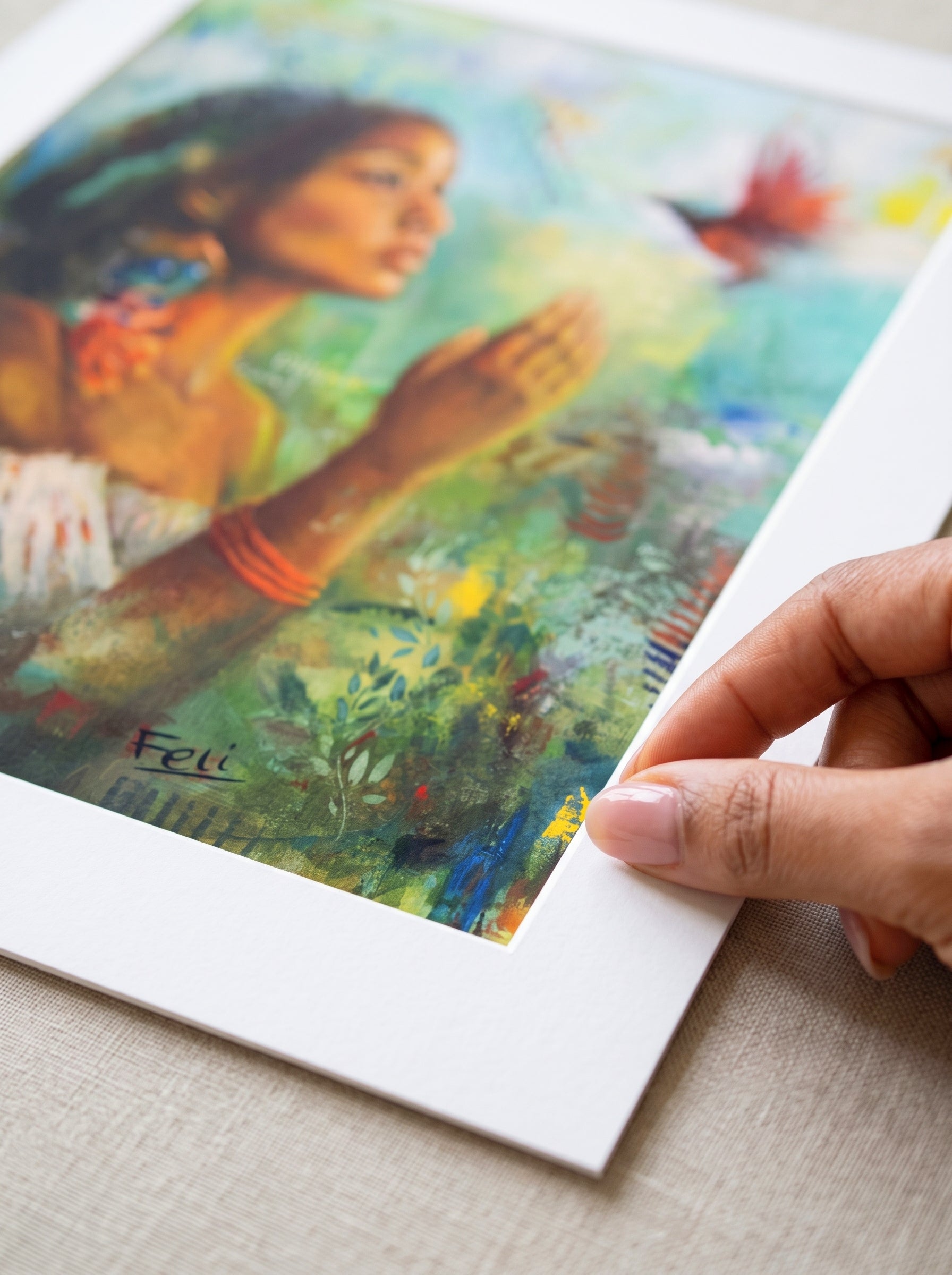 A close-up of a framed art print being held by a hand. The print features a colorful, textured painting of a black mixed indian woman reaching for a bird surrounded by green foliage  with a white mat border.
