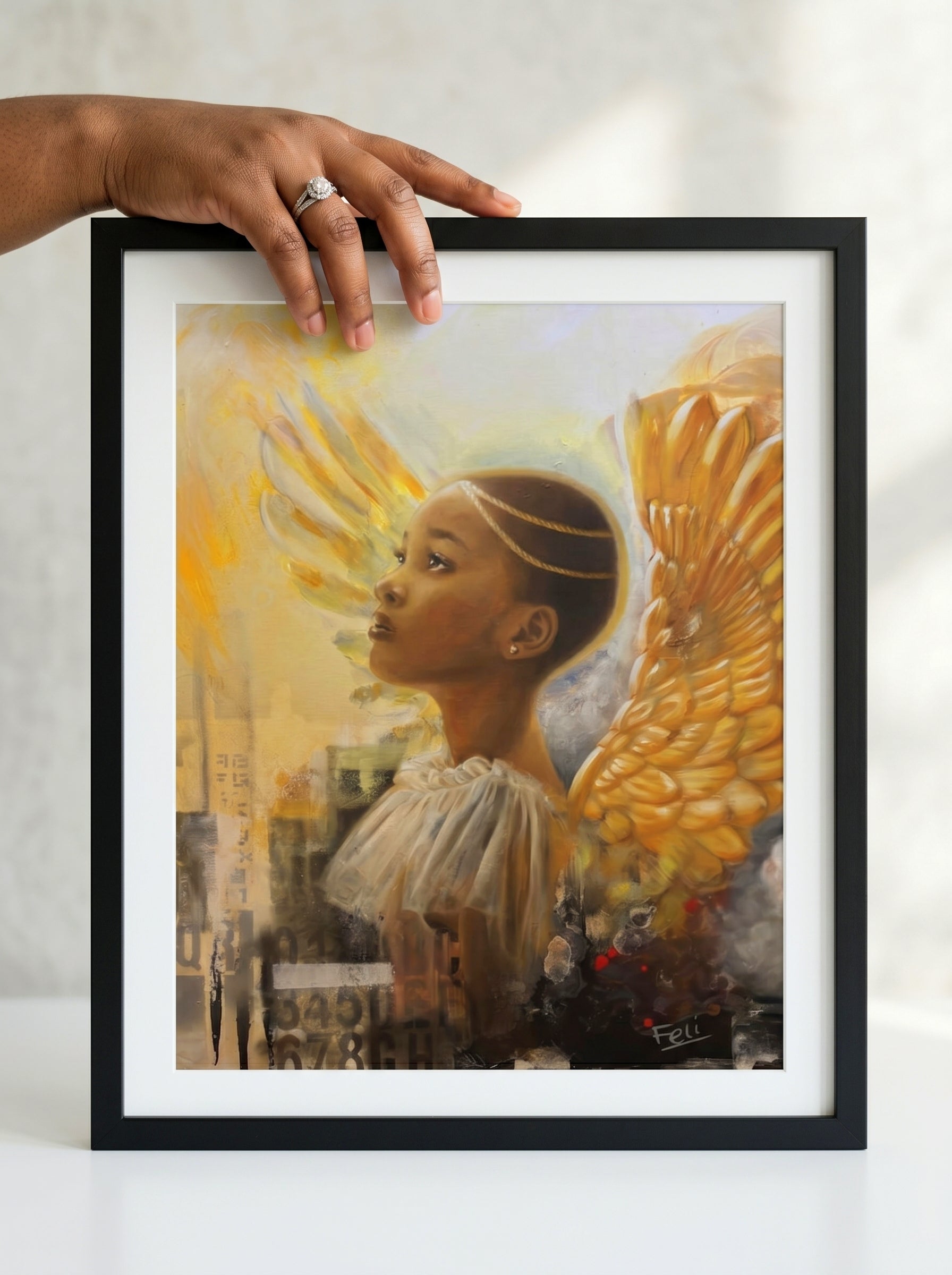 A framed art print entitled rays of hope by feli featuring a painted portrait of a black african girl woman with large golden angel wings in a black frame, held by a person's hand against a light neutral background.