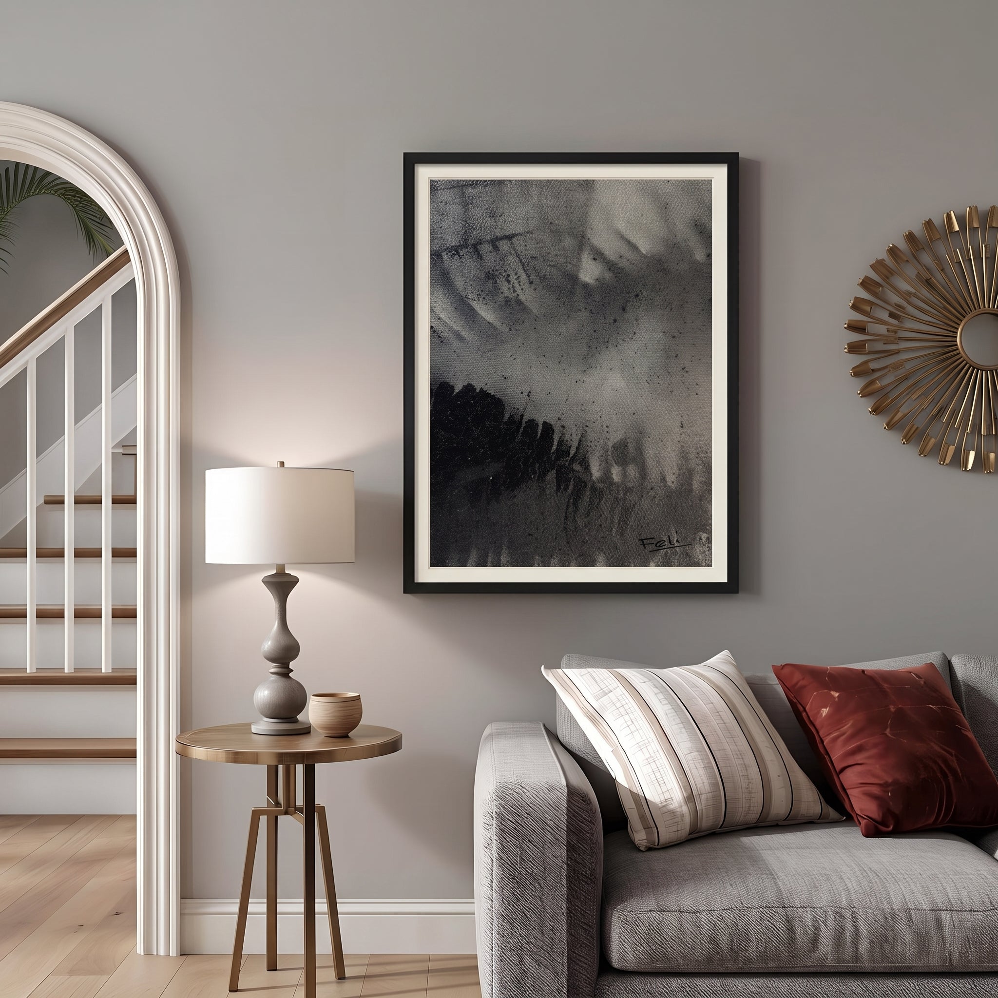 A framed abstract black and white art print entitled shadows of the past by feli the artist hangs in a black picture frame on a gray wall in a styled living room setting above a gray sofa with striped and red pillows, next to a wooden side table with a lamp.

