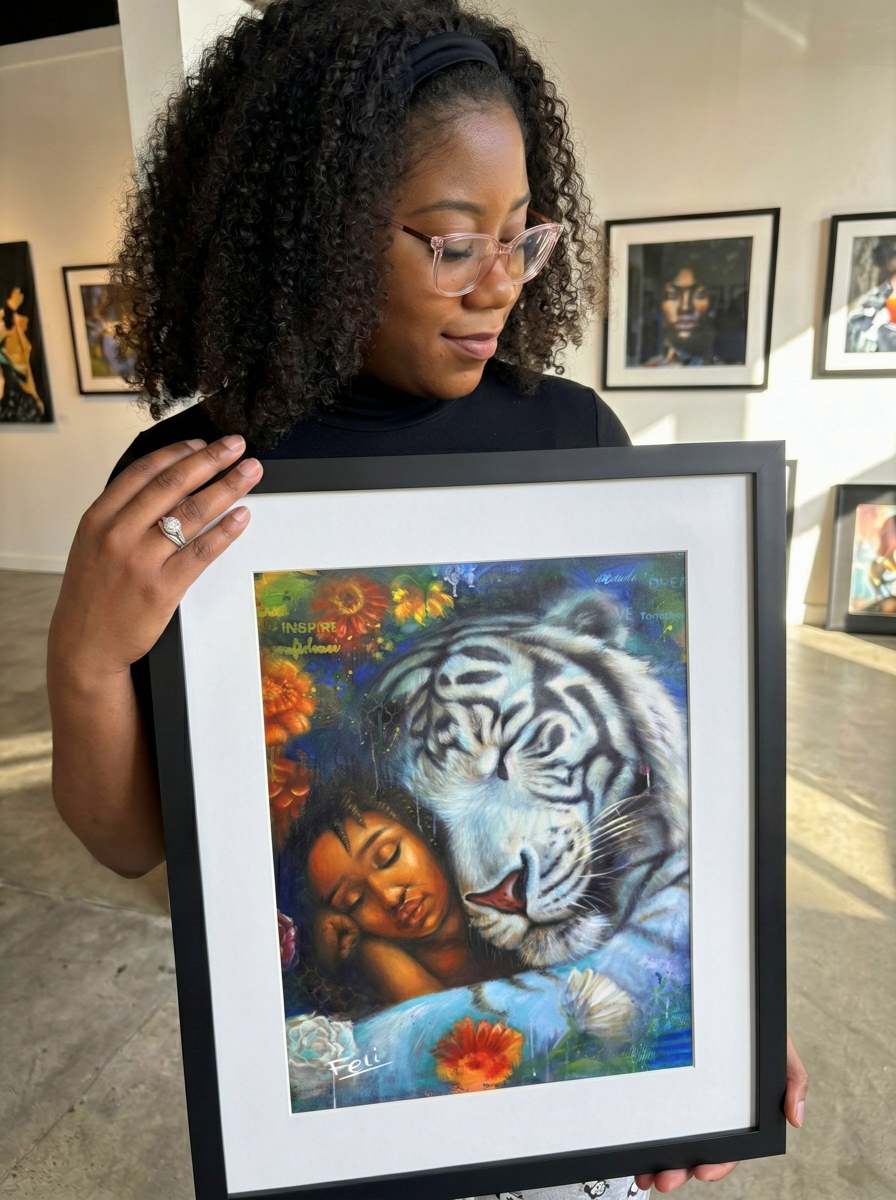A woman with curly hair and glasses holds up a framed art print entitled wild heart gentle soul by feli the artist featuring a white tiger and a woman with a blue hue with colorful flowers in the background. The art is framed in black with a white mat.