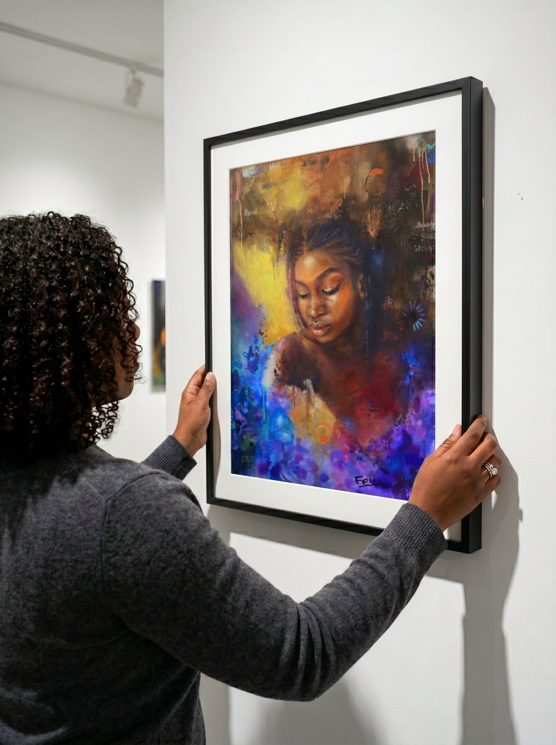 A person in a dark gray sweater hangs a framed colorful art print on a white gallery wall. The artwork shows a woman's face in an abstract, painterly style with blue, purple, yellow, and red tones, enclosed in a black frame with a white mat. The art print is entitled as she blooms by feli the artist.

